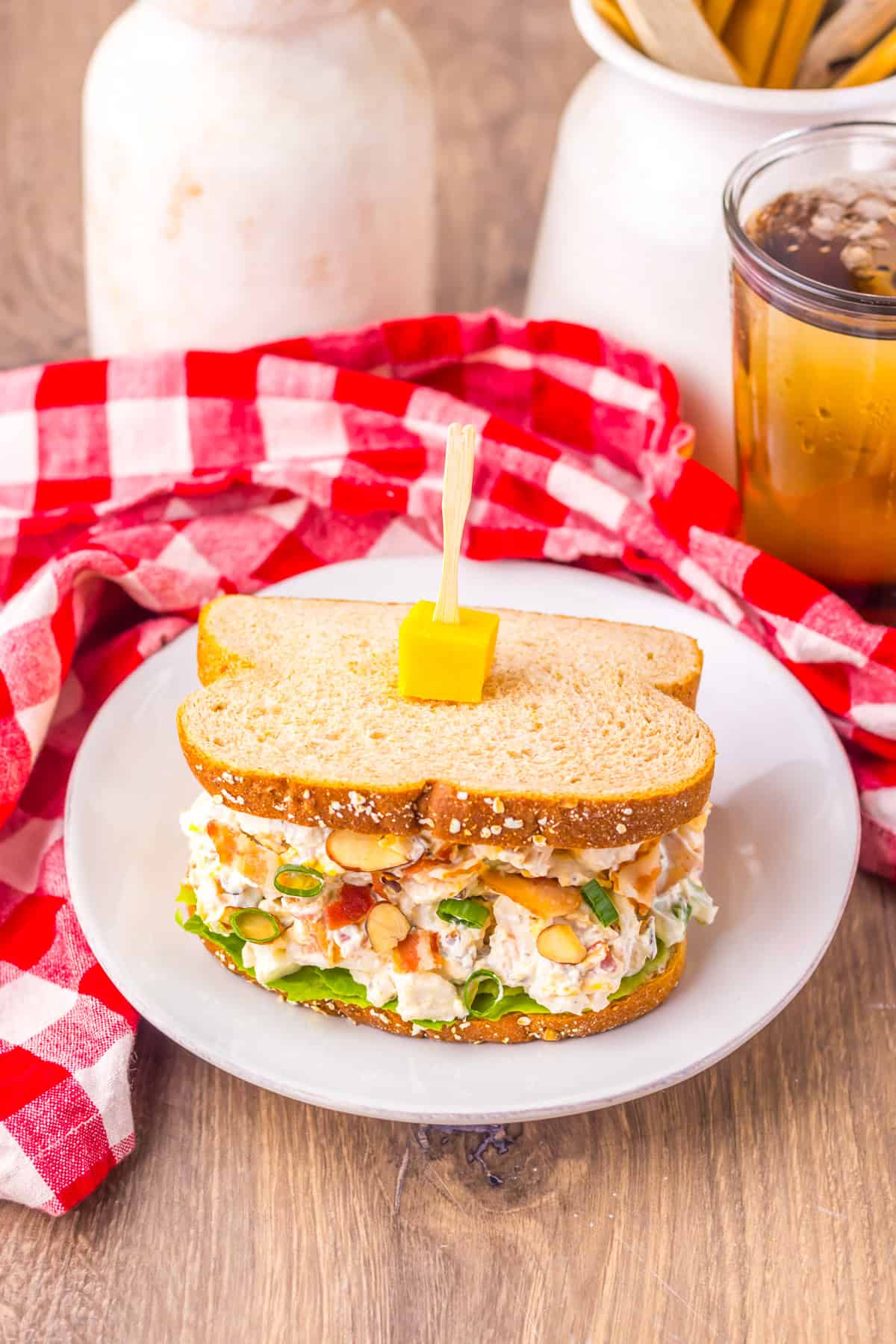 Top-down view of a chicken salad sandwich on a white plate, placed on a wooden table with a red checkered napkin and a glass of iced tea in the background.