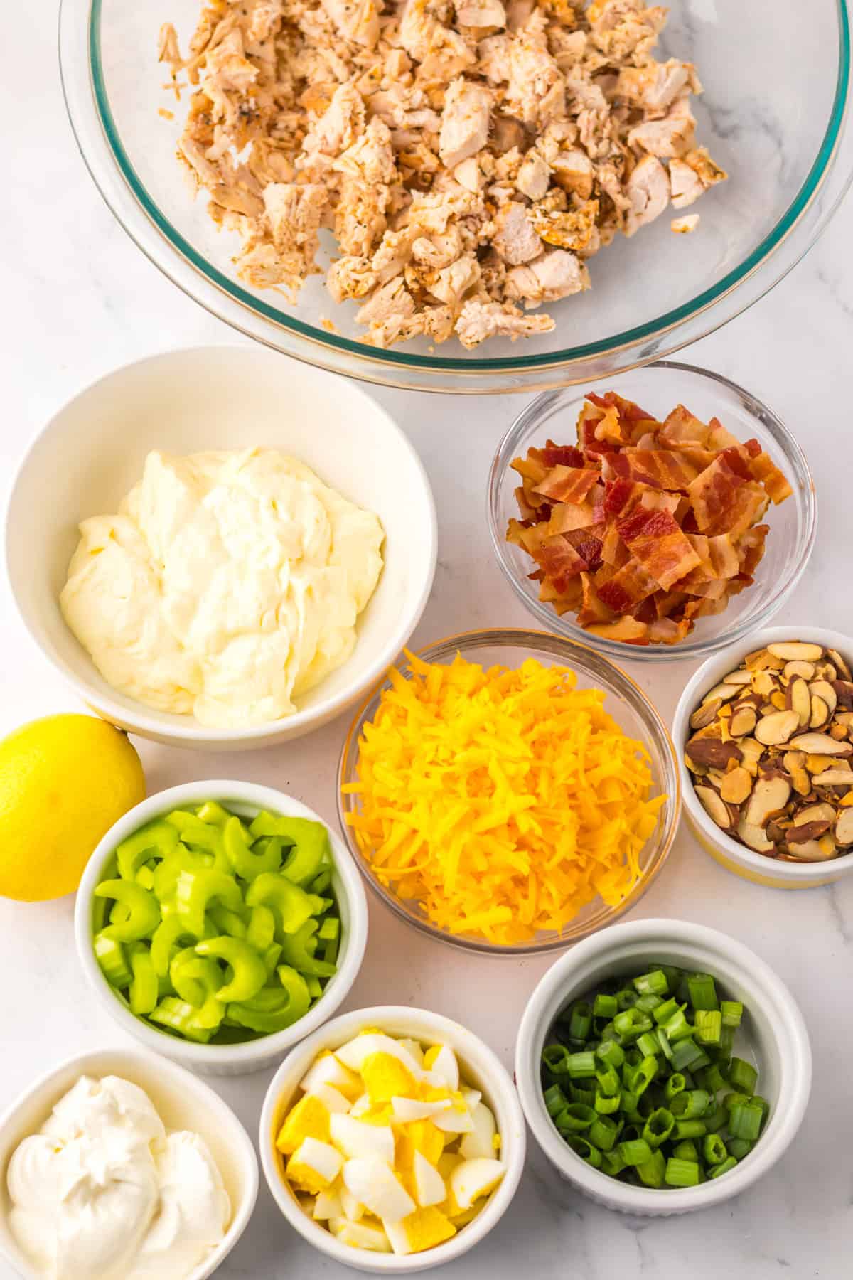 Flat lay of chicken salad ingredients including chopped chicken, mayonnaise, sour cream, cheddar, bacon, celery, hard-boiled eggs, green onions, sliced almonds, and a lemon.