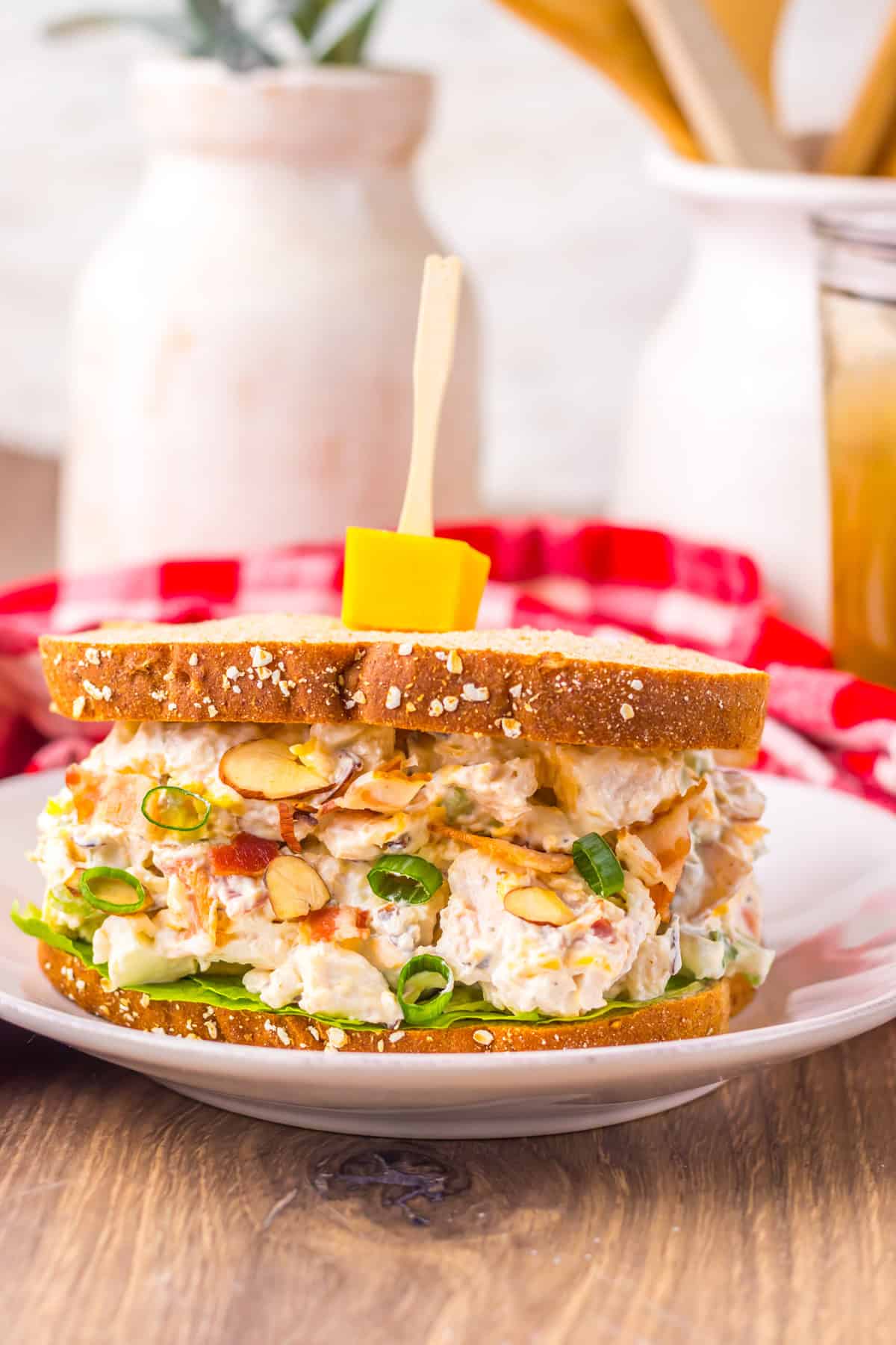 Rustic chicken salad sandwich on oat-topped bread, packed with bacon, green onions, almonds, and lettuce, served on a white plate with farmhouse kitchen decor in the background.