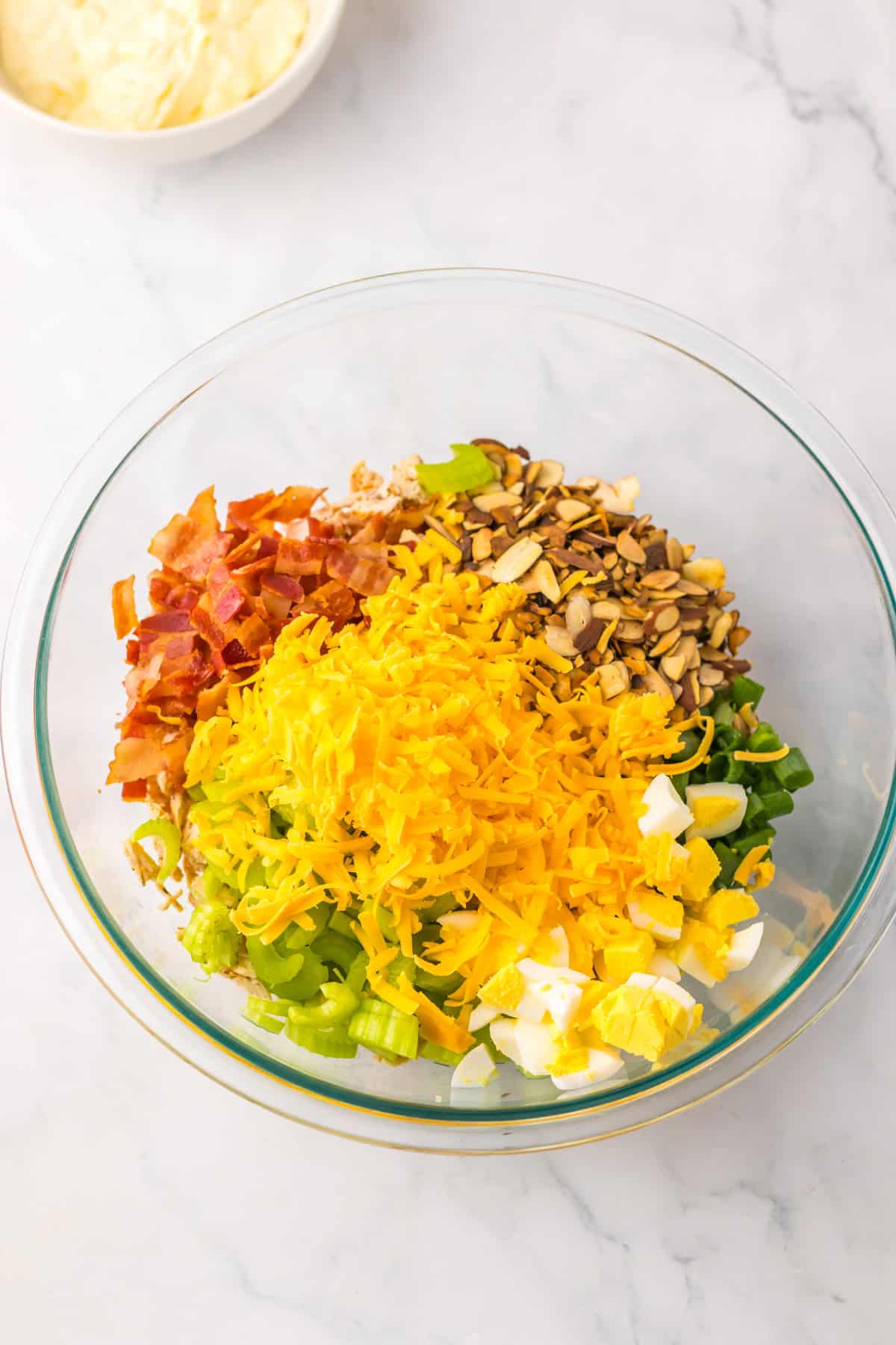 Bowl of ingredients for chicken salad including chopped bacon, shredded cheddar, celery, green onions, and chopped hard-boiled eggs, ready to be mixed with dressing.