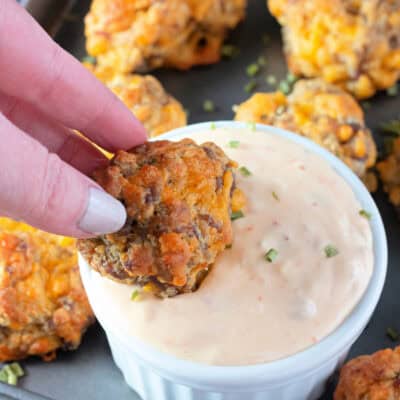 Close-up of a hand dipping a sausage ball into a creamy dipping sauce, with several sausage balls arranged in the background.