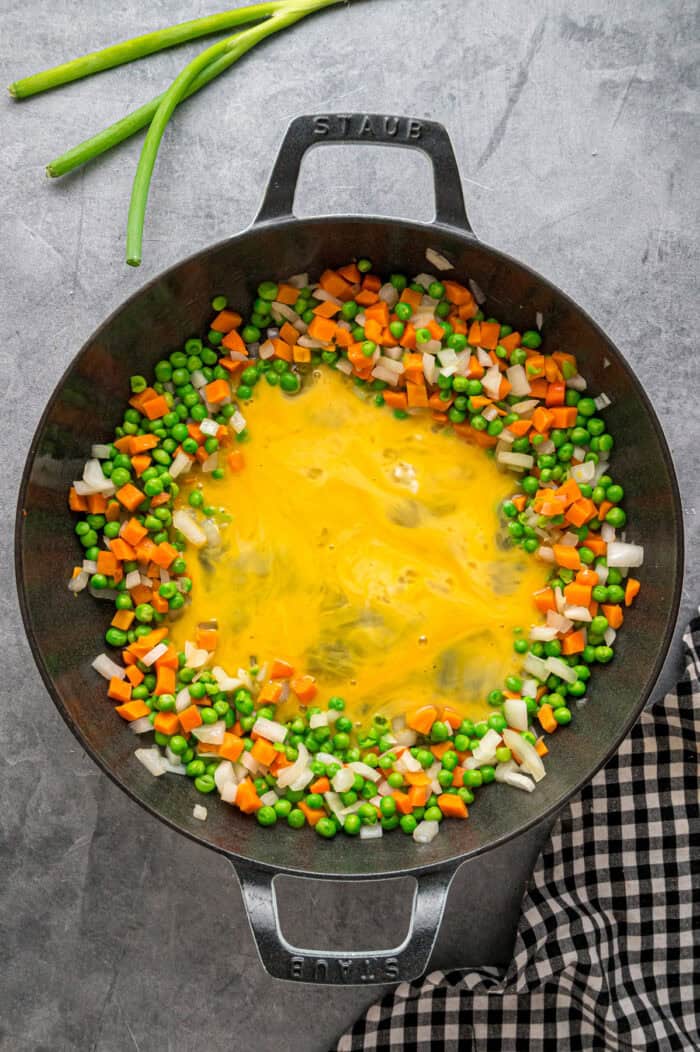 Scrambled eggs surrounded by onion, peas, and carrots in a wok